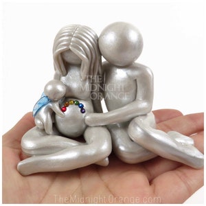 May include: A silver figurine of a couple holding a baby with a rainbow on the woman's belly. The baby has blue wings and is holding a small rainbow.