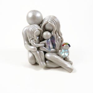 May include: A silver-colored family figurine depicting a parent and two children embracing. A small, rainbow-adorned figure sits nearby. The sculpture has a smooth, metallic finish and is set against a white background. The family is huddled together.