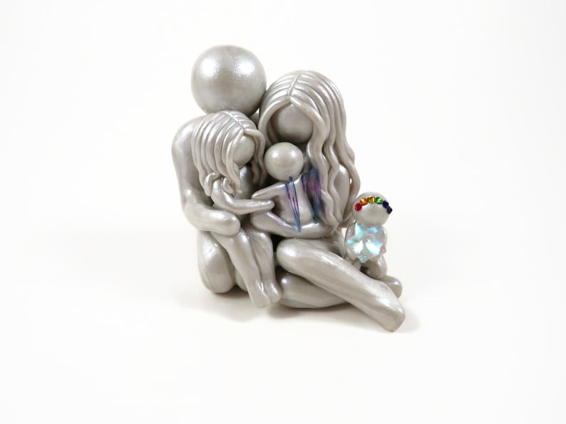 May include: A silver-colored family figurine depicting a parent and two children embracing. A small, rainbow-adorned figure sits nearby. The sculpture has a smooth, metallic finish and is set against a white background. The family is huddled together.