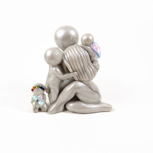 May include: A silver-colored sculpture of a mother figure embracing three children. One child has a rainbow-colored headband and iridescent clothing. Another child has blue and pink wings. The sculpture is on a white background.