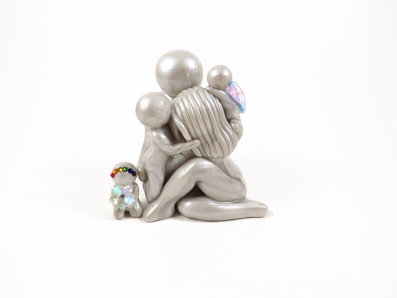May include: A silver-colored sculpture of a mother figure embracing three children. One child has a rainbow-colored headband and iridescent clothing. Another child has blue and pink wings. The sculpture is on a white background.