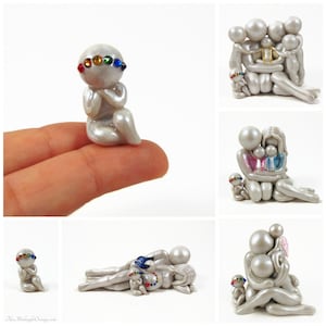 May include: A collection of handmade, pearl-like figurines. The figurines depict abstract human forms in various poses, some embracing, others in prayer. Each figure has colorful gemstone accents, with a rainbow of colors.