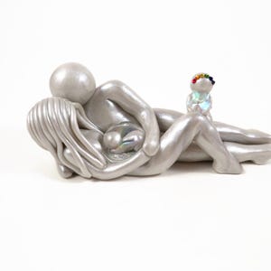 May include: A silver sculpture depicting a parent figure embracing another figure, with a smaller figure holding a rainbow-colored wreath. The figures are lying down, with a pearlescent orb in the center. The sculpture is on a white background.