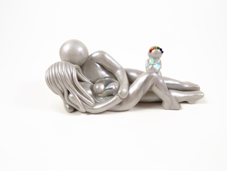 May include: A silver sculpture depicting a parent figure embracing another figure, with a smaller figure holding a rainbow-colored wreath. The figures are lying down, with a pearlescent orb in the center. The sculpture is on a white background.