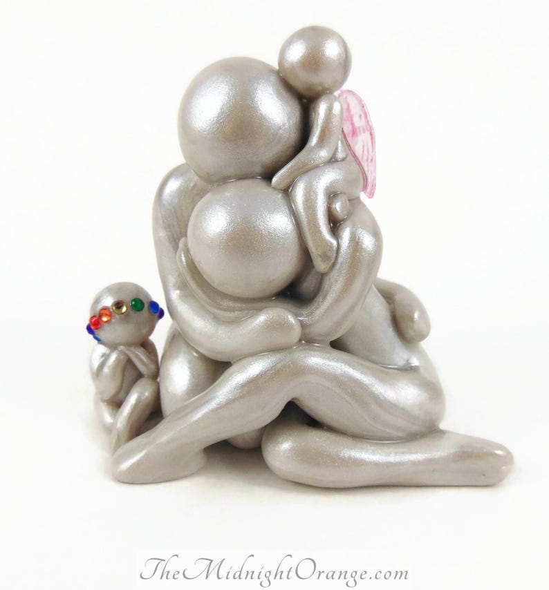 May include: A silver-colored sculpture depicting a family of four. The figures are stylized with rounded forms, with one child wearing a colorful beaded headband and another with pink wings. The Midnight Orange logo is at the bottom.
