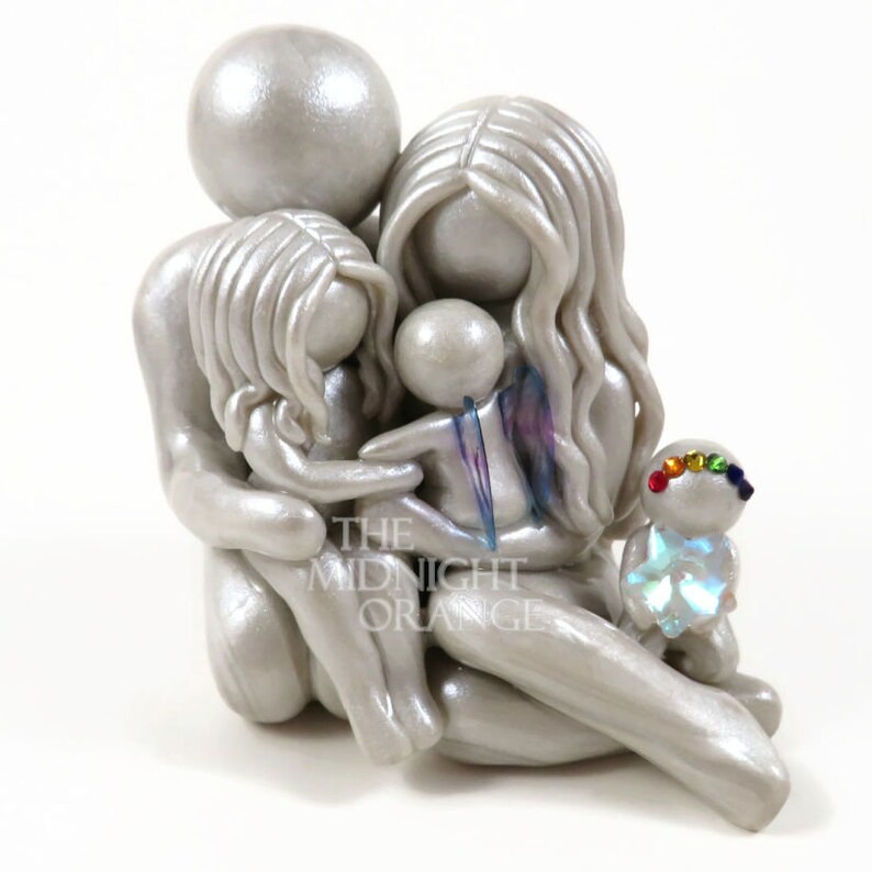 May include: A silver family figurine depicting a parent and two children embracing. A small figure with a rainbow-colored headpiece is also present. The figurine has a pearlescent finish and the text "THE MIDNIGHT ORANGE" is visible.