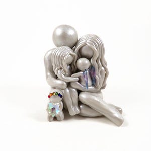 May include: A silver family figurine depicting a parent and two children embracing. A small figurine with a rainbow-colored headpiece stands in front. The family is seated, with the children nestled between the parents. The figurine is on a white background.
