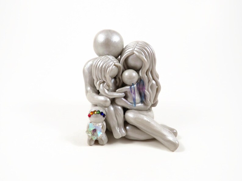 May include: A silver family figurine depicting a parent and two children embracing. A small figurine with a rainbow-colored headpiece stands in front. The family is seated, with the children nestled between the parents. The figurine is on a white background.
