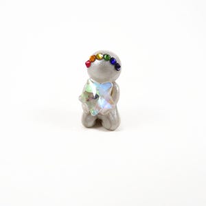 May include: A small, gray figurine in a kneeling pose, adorned with a rainbow of small gems on its head and a large, iridescent crystal in its arms. The figurine is set against a plain white background.
