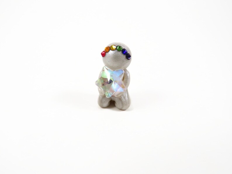 May include: A small, gray figurine in a kneeling pose, adorned with a rainbow of small gems on its head and a large, iridescent crystal in its arms. The figurine is set against a plain white background.