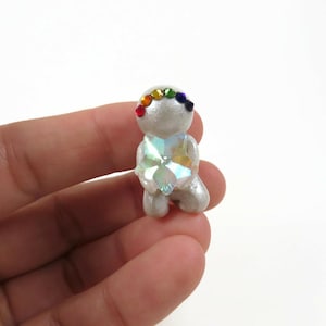 May include: A small, silver-colored figurine in a kneeling pose, adorned with a rainbow-colored gemstone headband. The figurine has a large, multi-faceted crystal heart. The figurine is held in a person's hand.