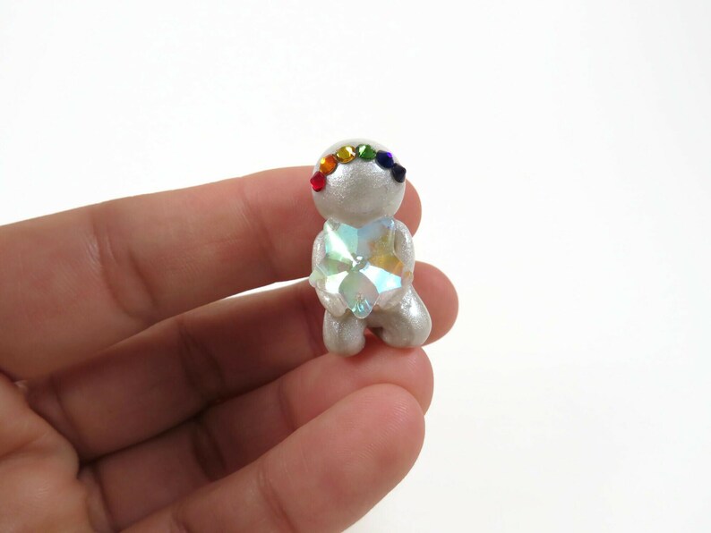 May include: A small, silver-colored figurine in a kneeling pose, adorned with a rainbow-colored gemstone headband. The figurine has a large, multi-faceted crystal heart. The figurine is held in a person's hand.