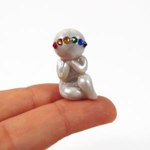 May include: A small, pearlescent figurine of a seated figure with hands clasped in prayer. The figure has a rainbow-colored gemstone band across its forehead. The figurine is held between two fingers.