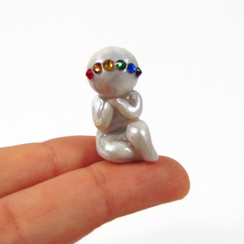 May include: A small, pearlescent figurine of a seated figure with hands clasped in prayer. The figure has a rainbow-colored gemstone band across its forehead. The figurine is held between two fingers.