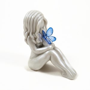 Faceless figurine of a mother sitting with knees bent upright and holding a baby on her knees, cradled to her chest. The baby is  wearing bright blue butterfly wings. The hair and wings are customizable for this sculpture.