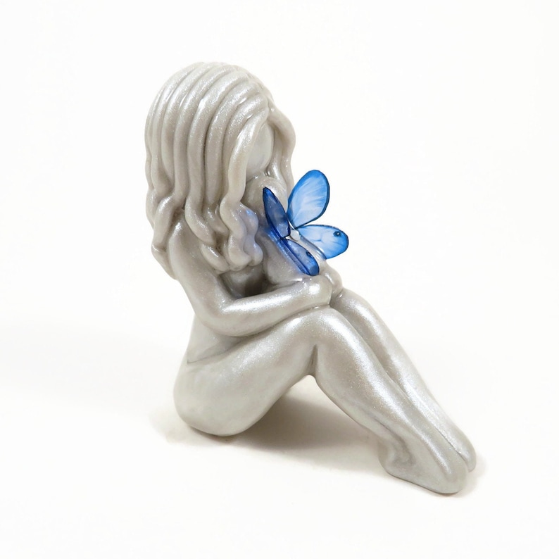 Faceless figurine of a mother sitting with knees bent upright and holding a baby on her knees, cradled to her chest. The baby is  wearing bright blue butterfly wings. The hair and wings are customizable for this sculpture.