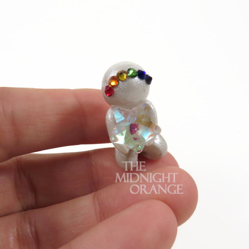 May include: A small, silver figurine with a rainbow-colored gemstone headband and a large, iridescent star held in its hands. The figurine is detailed with a pearlescent finish. The text "THE MIDNIGHT ORANGE" is visible on the base.