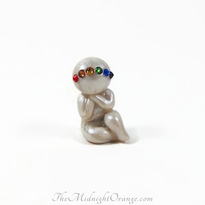 May include: A small, pearlescent figurine of a seated figure with a rainbow of colored gemstones across the forehead. The figure is in a meditative pose with hands clasped. The Midnight Orange.com is printed at the bottom.