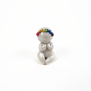 May include: A small, silver figurine of a person in a seated position with hands clasped in prayer. The figurine has a rainbow-colored gemstone crown. The figure is on a white background.