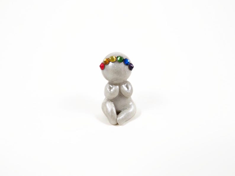 May include: A small, silver figurine of a person in a seated position with hands clasped in prayer. The figurine has a rainbow-colored gemstone crown. The figure is on a white background.