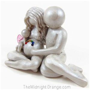 May include: A white ceramic figurine of a pregnant woman and a man embracing. The woman's belly is decorated with a rainbow of rhinestones. The man has his arm around the woman's shoulder. The figurine is a symbol of love, family, and hope.