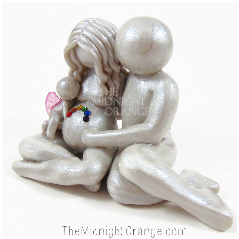 May include: A white ceramic figurine of a pregnant woman and a man embracing. The woman's belly is decorated with a rainbow of rhinestones. The man has his arm around the woman's shoulder. The figurine is a symbol of love, family, and hope.