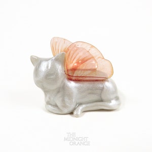 Clay minimalistic cat sculpture in a pearl color. Kitty is in laying pose with its head up and wearing pastel orange butterfly wings.