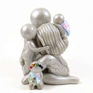 May include: A silver figurine depicting a parent figure embracing three children. One child has a rainbow-colored headband and a crystal-like chest. The figurine has a pearlescent finish. The Midnight Orange is printed on the base.
