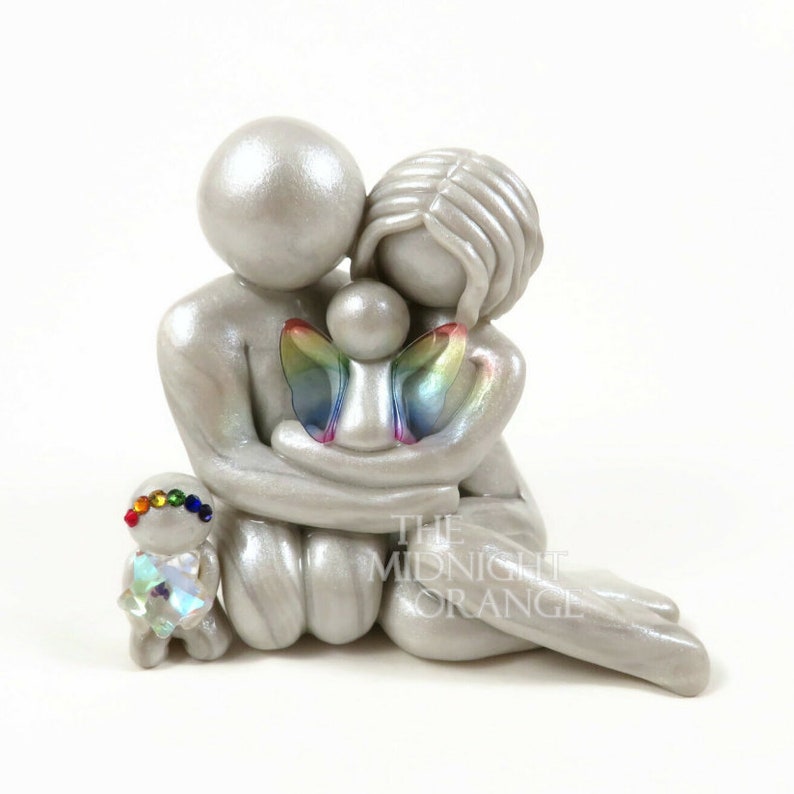 May include: A silver figurine depicting a family of four. The parents embrace a child with rainbow wings. A smaller figure with a rainbow-colored head and a crystal heart kneels nearby. The Midnight Orange is printed on the bottom right.