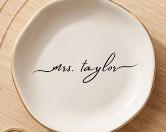 May include: A round, off-white ceramic trinket dish with an irregular, organic edge and a gold-painted rim. The center features the black cursive text "mrs. taylor". The dish is accompanied by a gold chain necklace with a pearl pendant and a gold bracelet.