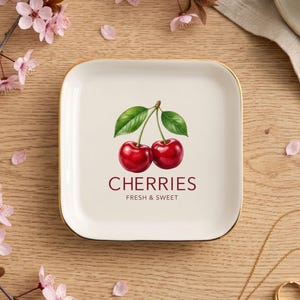 May include: A square white ceramic trinket dish with rounded corners and a gold-toned rim. The center features a detailed illustration of two red cherries with green leaves, accompanied by the text CHERRIES and FRESH & SWEET in dark red.