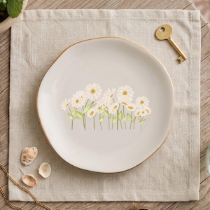 May include: A round, off-white ceramic trinket dish featuring a delicate illustration of white daisies with yellow centers and green stems. The dish has a smooth, slightly irregular edge finished with a thin, metallic gold-colored rim.
