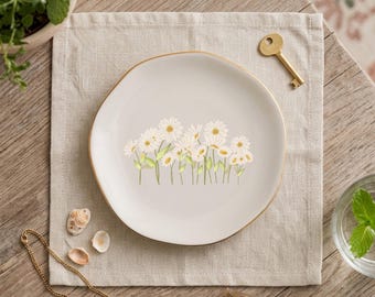 May include: A round, off-white ceramic trinket dish featuring a delicate illustration of white daisies with yellow centers and green stems. The dish has a smooth, slightly irregular edge finished with a thin, metallic gold-colored rim.