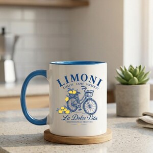 May include: A white ceramic mug with a blue handle and interior rim. The front features a blue graphic of a bicycle with a basket of yellow lemons, accompanied by the text LIMONI, AMALFI CAPRI SORRENTO, La Dolce Vita, and MEDITERRANEAN TRADITION.
