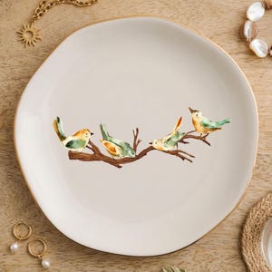 May include: A round, off-white ceramic plate featuring a central illustration of four small birds with yellow and green plumage perched on a brown tree branch. The plate has a smooth, slightly irregular edge and a thin gold-colored rim.
