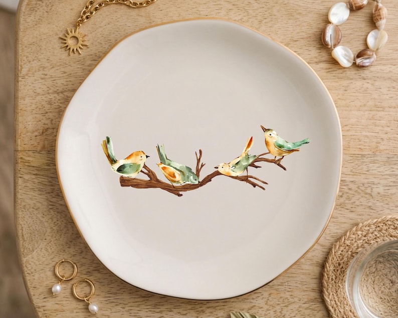 May include: A round, off-white ceramic plate featuring a central illustration of four small birds with yellow and green plumage perched on a brown tree branch. The plate has a smooth, slightly irregular edge and a thin gold-colored rim.