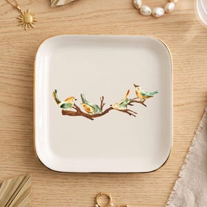 May include: A square white ceramic trinket dish with rounded corners and a thin gold-toned rim. The center of the dish features a watercolor illustration of four small birds perched on a brown tree branch with green and orange accents.