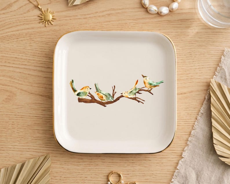 May include: A square white ceramic trinket dish with rounded corners and a thin gold-toned rim. The center of the dish features a watercolor illustration of four small birds perched on a brown tree branch with green and orange accents.
