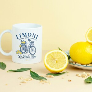 May include: A white ceramic mug featuring a blue graphic of a bicycle with a basket of lemons, the text LIMONI AMALFI CAPRI SORRENTO, and La Dolce Vita MEDITERRANEAN TRADITION. Beside the mug is a plate with whole and halved yellow lemons.
