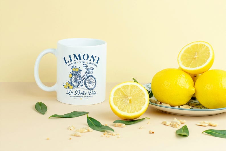 May include: A white ceramic mug featuring a blue graphic of a bicycle with a basket of lemons, the text LIMONI AMALFI CAPRI SORRENTO, and La Dolce Vita MEDITERRANEAN TRADITION. Beside the mug is a plate with whole and halved yellow lemons.