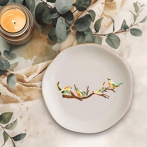 May include: A round, off-white ceramic dish featuring a watercolor-style illustration of four small birds perched on a brown tree branch. The dish has a smooth, slightly irregular edge and a clean, neutral surface, providing a simple display.