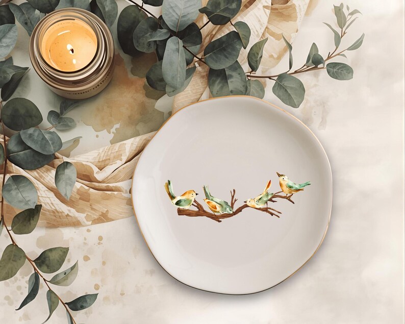 May include: A round, off-white ceramic dish featuring a watercolor-style illustration of four small birds perched on a brown tree branch. The dish has a smooth, slightly irregular edge and a clean, neutral surface, providing a simple display.