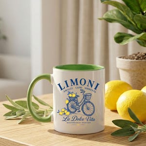May include: A white ceramic mug with a green interior and handle, featuring a blue graphic of a bicycle with a basket of lemons. The text reads LIMONI AMALFI CAPRI SORRENTO and La Dolce Vita MEDITERRANEAN TRADITION in blue lettering on the front.
