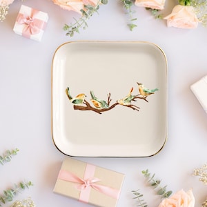 May include: A square ceramic trinket dish with rounded corners and a gold-toned rim. The center of the off-white dish features a watercolor illustration of four small birds perched on a brown tree branch with green and orange plumage details.
