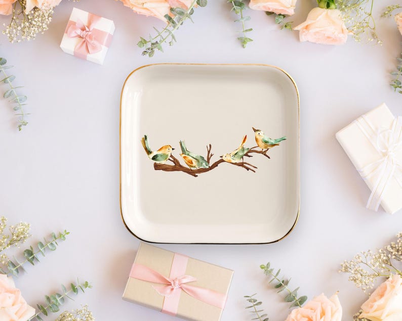 May include: A square ceramic trinket dish with rounded corners and a gold-toned rim. The center of the off-white dish features a watercolor illustration of four small birds perched on a brown tree branch with green and orange plumage details.