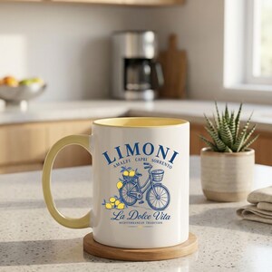 May include: A white ceramic mug with a yellow interior and handle, featuring a blue graphic of a bicycle carrying lemons in a basket. The text reads LIMONI AMALFI CAPRI SORRENTO and La Dolce Vita MEDITERRANEAN TRADITION in blue and yellow lettering.