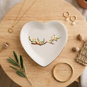 May include: A white heart-shaped ceramic trinket dish featuring a central illustration of four small birds perched on a brown branch. The dish has a thin gold-colored rim and is surrounded by a gold necklace, gold hoop earrings, and a gold bangle.