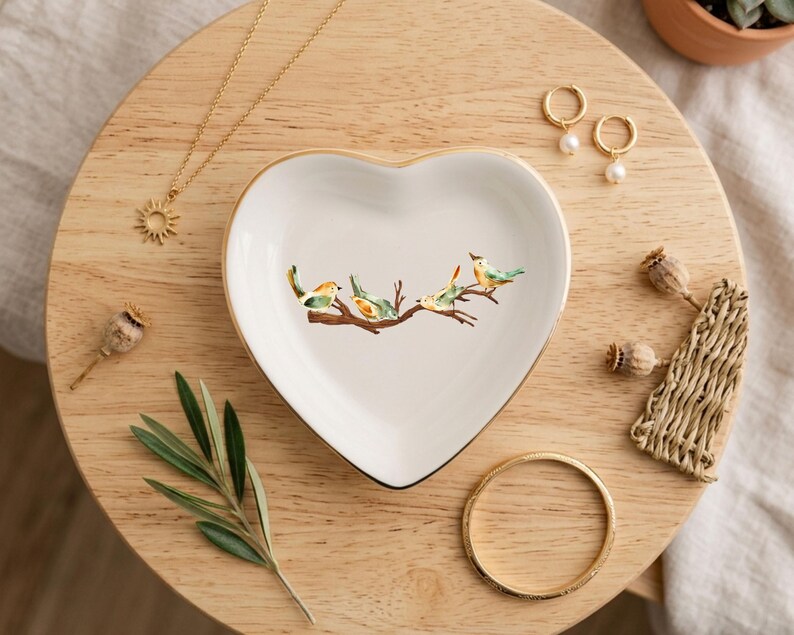 May include: A white heart-shaped ceramic trinket dish featuring a central illustration of four small birds perched on a brown branch. The dish has a thin gold-colored rim and is surrounded by a gold necklace, gold hoop earrings, and a gold bangle.