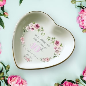 May include: A heart-shaped ceramic dish with a gold rim, featuring floral designs and text that reads "Ruby Wedding Anniversary 40th". The dish is surrounded by pink peonies on a light blue background.