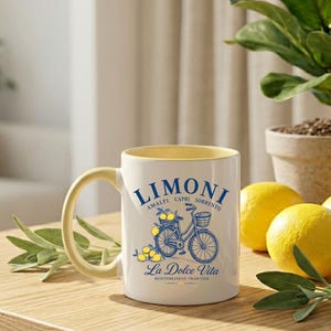 May include: A white ceramic mug with a pale yellow interior and handle. The front features a blue graphic of a bicycle with a basket of lemons, surrounded by the text LIMONI, AMALFI CAPRI SORRENTO, and La Dolce Vita MEDITERRANEAN TRADITION.